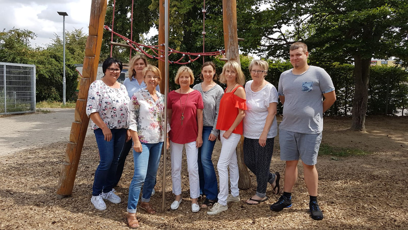 Acht Erwachsene stehen neben einem Holzkletter Gerüst auf einem Spiel Platz und lächeln freundlich in die Kamera