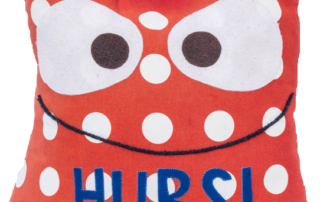 Ein weiches rotes Stoff-Tier mit großen Augen weißen Punkten blauen Beinen und dem Wort HUBSI auf der Brust das Maskottchen der Schule freigestellt