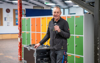 Hausmeister steht lächelnd mit Mikrofon an der Soundbox vor den bunten Spinten im Flur 2