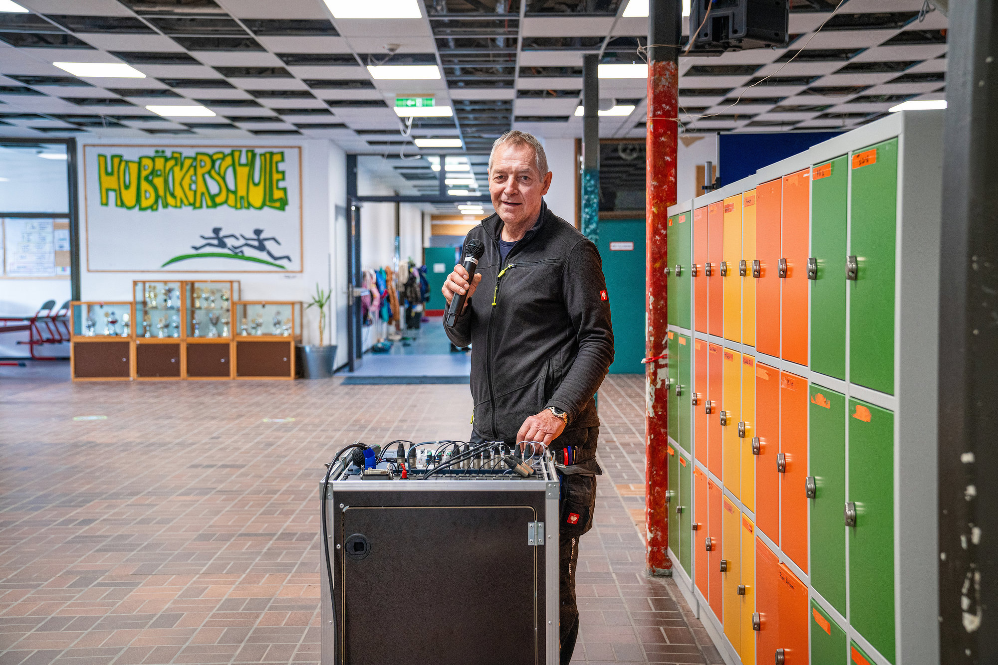 Hausmeister steht lächelnd mit Mikrofon an der Soundbox vor den bunten Spinten im Flur