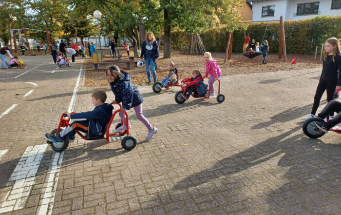 Mehrere Kinder fahren mit roten Dreirädern auf dem Schul Hof und haben Spass