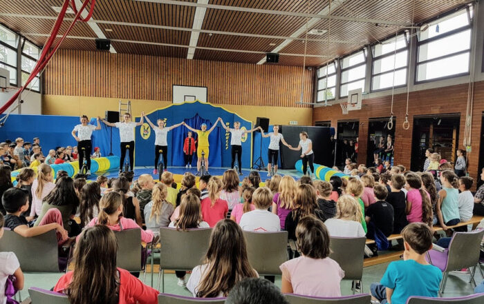 Mehrere Künstler stehen auf einer Zirkus Bühne auf Einrädern vor vielen Kindern die fröhlich zusehen und halten sich an der Hand