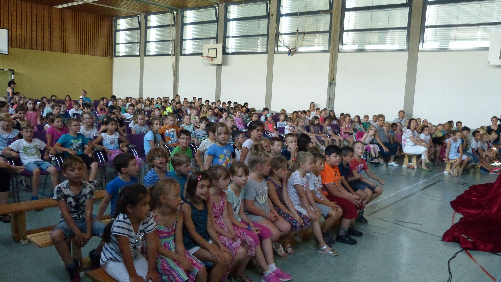 Viele Kinder sitzen auf Bänken in einer großen Halle und schauen nach vorne